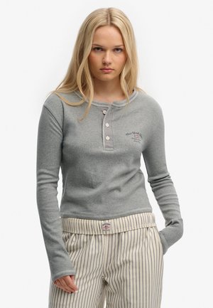 Joven mujer con cabello largo y rubio, vestida con una blusa gris de manga larga abotonada y pantalones beige a rayas con una cinturilla con logo.