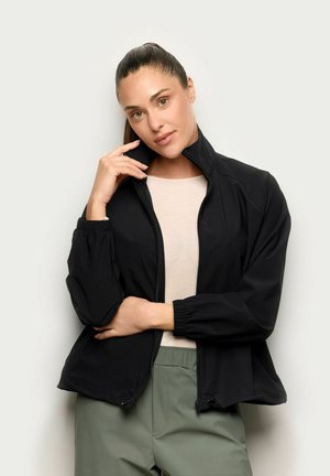 Kobieta z kitką, ubrana w czarną kurtkę na zamek błyskawiczny na jasną koszulę i zielone spodnie, pozująca z ręką przy twarzy na jednolitym tle.