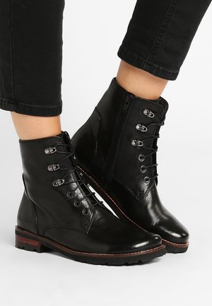 Bottes chevilles en cuir noir avec lacets sur le devant, fermetures éclair latérales et petits talons empilés, portées avec un pantalon noir.