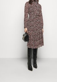 Mujer vistiendo un vestido floral negro de manga larga con cinturón, botas negras hasta la rodilla y sosteniendo un bolso negro frente a un fondo liso.