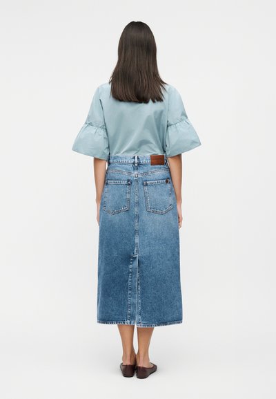 Γυναίκα με ίσια σκούρα μαλλιά που φοράει ανοιχτό μπλε μπλούζα με κοντά μανίκια και φούστα μεσαίου μήκους από μπλε τζιν με ψηλή μέση, εμφανιζόμενη από πίσω.
