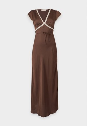 Femme portant une longue robe marron avec bordure blanche, des talons noirs, et tenant un sac à main blanc tissé sur un fond clair uni.