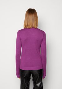 Femme aux cheveux raides de longueur moyenne portant un pull violet à manches longues et un pantalon en cuir noir, debout face à un mur blanc uni.