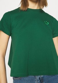 T-shirt en coton vert avec un col rond et des manches courtes. Présente un logo brodé sur la poitrine. Texture douce et coupe décontractée.