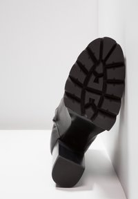 Bottine en cuir noir avec un talon épais et une semelle en caoutchouc texturée dotée d'une profonde bande de roulement pour une meilleure adhérence. L'angle met en valeur la semelle et le design du talon.