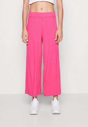 Femme portant un pantalon large rose vif et des baskets blanches, debout devant un fond blanc uni.
