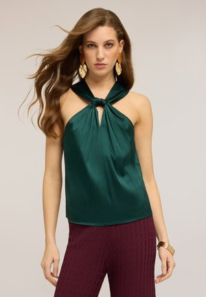 Femme aux cheveux longs et ondulés portant un haut dos-nu vert, un pantalon bordeaux texturé, de grandes boucles d'oreilles dorées et ambrées, ainsi que des bracelets dorés au poignet droit.