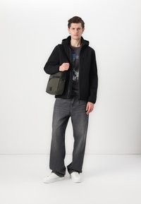 Casaco preto com fecho éclair e capuz, t-shirt gráfica por baixo, jeans largos cinzentos, sapatilhas brancas e mala transversal em tom oliva.