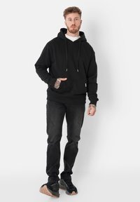 Svart hettegenser med frontlomme, snøring ved hetta og ribbestrikkede mansjetter, kombinert med slitte svarte jeans og svarte sneakers.
