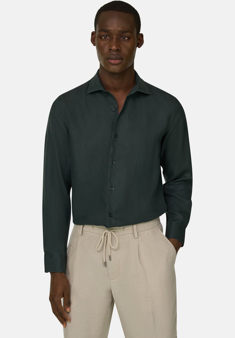 Man draagt een donkergroen overhemd met lange mouwen, ingestopt in beige broek met trekkoord, staat met één hand in zijn zak.