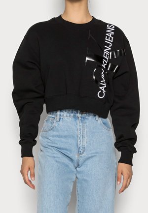 Personne portant un sweat court noir avec le texte "Calvin Klein Jeans" et un détail de ruban noir, associé à un jean taille haute bleu clair.