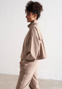 Béžová zkrácená bunda vyrobená z lehkého materiálu, s vysokým límcem, kapsou na zip a volnými rukávy, doplněná o ladící kalhoty.