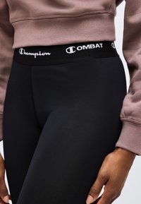 Personne portant des leggings noirs Champion Combat et un sweat court mauve, main reposant près de la cuisse, taille et haut de la jambe visibles.