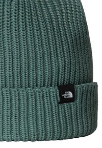 Beanie verde lavorato a maglia con texture a coste, dotato di una toppa nera sul polsino. Il materiale sembra morbido ed elastico.
