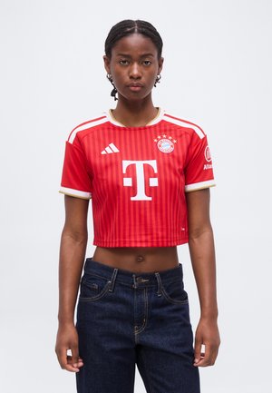 Vrouw draagt een rood kort Bayern München voetbalshirt met logo's, staand tegen een effen lichte achtergrond, vooruit kijkend.