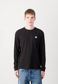 Zwarte lange mouw geribbelde t-shirt met een ronde halslijn en een kleine logo patch op de borst, gecombineerd met lichtblauwe denim jeans.