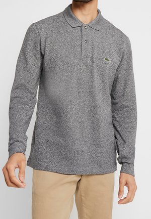 Mann trägt ein graues langärmeliges Poloshirt mit Knöpfen und beige Hose, steht vor einem schlichten Hintergrund.