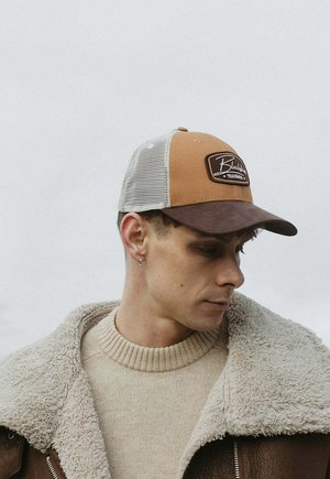 Young man wearing beige sweater, brown and tan mesh cap with patch, and brown shearling jacket, looking downward against plain background.