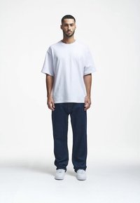Λευκό μεγάλο βαμβακερό T-shirt φορεμένο με σκούρες μπλε τζην παντελόνες και λευκά αθλητικά παπούτσια, σε απλό, ανοιχτό φόντο.