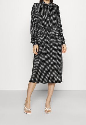 Femme portant une robe midi noire à manches longues avec des pois blancs, associée à des sandales blanches à talons et à brides.