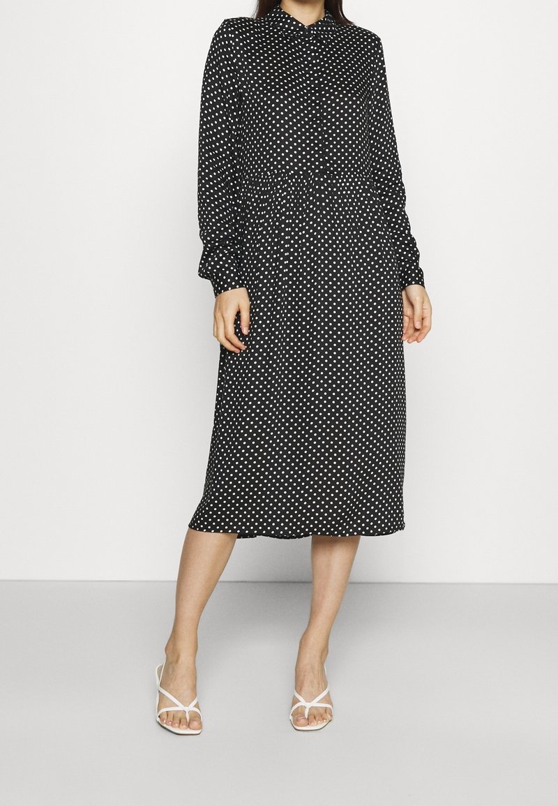 Femme portant une robe midi noire à manches longues avec des pois blancs, associée à des sandales blanches à talons et à brides.