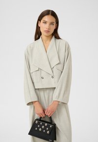 Donna con lunghi capelli castani che indossa un blazer oversize grigio chiaro e pantaloni, tiene una borsa nera con borchie argentate davanti a uno sfondo semplice.