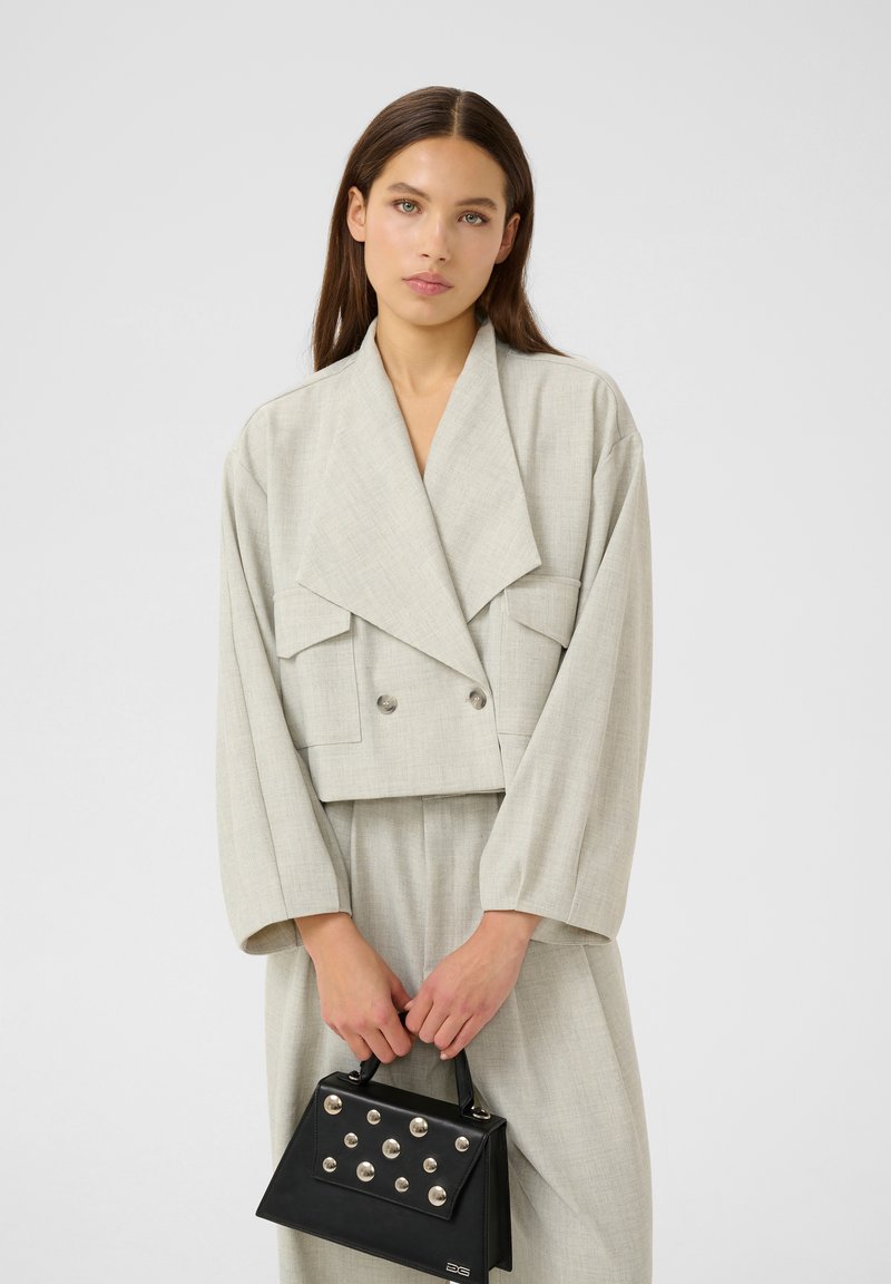 Donna con lunghi capelli castani che indossa un blazer oversize grigio chiaro e pantaloni, tiene una borsa nera con borchie argentate davanti a uno sfondo semplice.