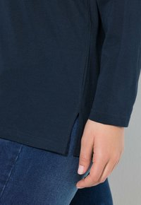 Dunkelblaues Langarmshirt aus Baumwolle mit seitlichen Schlitzen am Saum, kombiniert mit dunklen Jeans, wobei eine Hand auf dem Oberschenkel ruht.