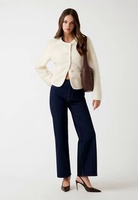 Veste crème texturée à boutons, col arrondi. Associée à un jean large bleu foncé et des chaussures à talons marron.