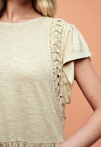 Helles beige Kurzarmtop mit einer Spitzenauflage auf den Schultern, sanfter Textur und gerafftem Saum.