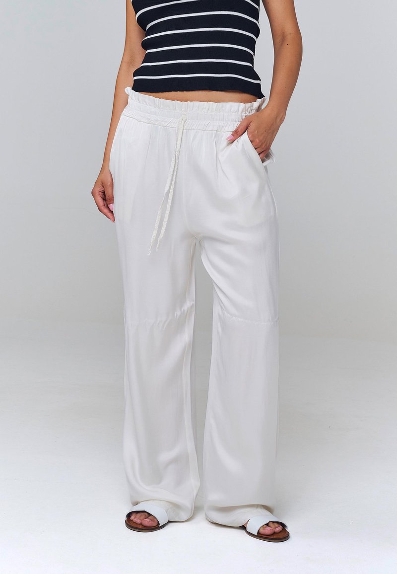 Femme portant un pantalon blanc taille haute à jambes larges avec cordon de serrage et poches, associé à un top sans manches rayé noir et blanc et des sandales blanches.