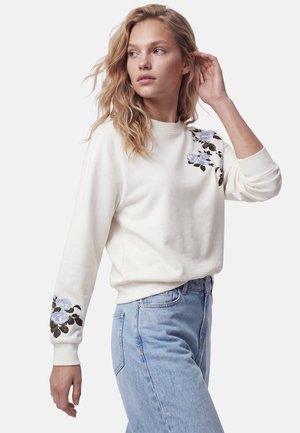 Jeune femme portant un sweat-shirt blanc avec une broderie florale bleue sur l'épaule et la manche, associée à un jean bleu clair, regardant sur le côté.