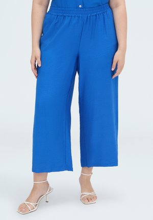 Persoon die felblauwe, wijde cropped broek draagt met elastische tailleband en witte sandalen met hoge hak en bandjes, staand op een effen achtergrond.