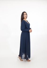 Vrouw met lang donker haar, gekleed in een navy lange mouw maxi-jurk en sandalen met transparante hakken, staand tegen een witte achtergrond.