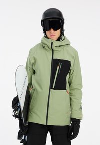 Giacca da snowboard resistente all'acqua di colore verde con dettagli neri, tasche con zip e cappuccio regolabile. Indossata con un casco nero e mentre si tiene una tavola da snowboard.