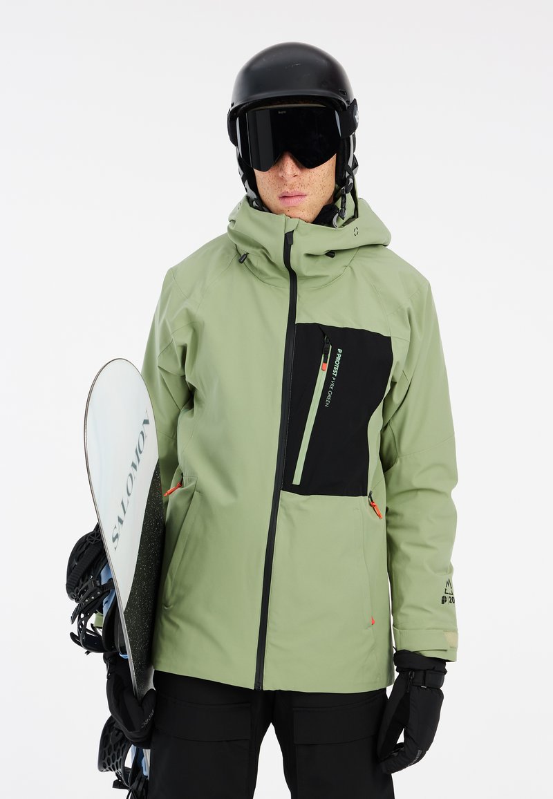 Giacca da snowboard resistente all'acqua di colore verde con dettagli neri, tasche con zip e cappuccio regolabile. Indossata con un casco nero e mentre si tiene una tavola da snowboard.