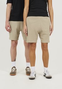 Shorts beige côtelés avec un motif texturé, affichant une coupe décontractée. Associés à des chemises noires et à divers types de chaussures, présentés ensemble.