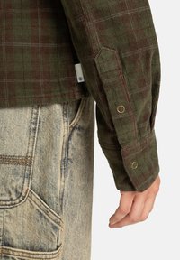 Mano in posizione rilassata accanto a pantaloni in denim blu sbiadito con tasche a toppa e maniche di una camicia di côtone a quadri verdi usurata con bottoni a pressione metallici.
