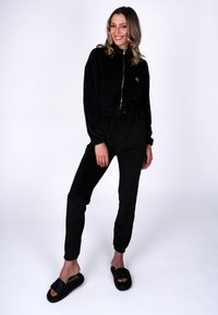 Vrouw die lacht, staand, gekleed in een zwarte jumpsuit met rits en elastische boorden, en zwarte slippers, tegen een witte achtergrond.