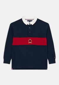 Polo de manga larga en color navy con cuello blanco, franja horizontal roja y emblema bordado. Cuenta con una tapeta de tres botones y puños acanalados.
