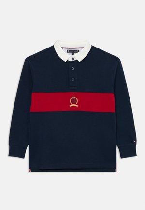Polo-shirt à manches longues bleu marine avec un col blanc, une rayure horizontale rouge et un emblème brodé. Dispose d'une patte de boutonnage à trois boutons et de poignets à nervures.