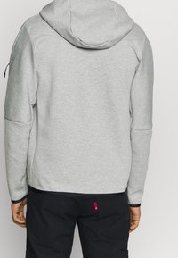 Sudadera con capucha gris, de manga larga, con una textura suave y un dobladillo en navy que contrasta. Cuenta con un bolsillo con cremallera en la manga.