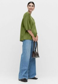 Överdimensionerad grön topp med korta ärmar, kombinerad med vida ljusblå jeans. Modellen håller en liten brun handväska och har på sig svarta skor.