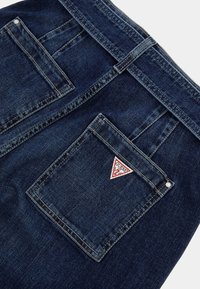 Tamsiai mėlynos džinsinės kelnės su galiniu kišeniu, kuriame yra trikampis raudonai baltas logotipas, siuvimo detalės ir klasikinių juosmenų dizainas.