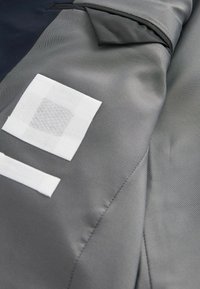 Una tela gris texturizada presenta dos parches blancos, uno cuadrado y uno rectangular, con un borde suave y una sección superior de color contrastante.