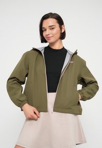 Γυναίκα με κοντά σκούρα μαλλιά φοράει ένα χακί μπουφάν με φερμουάρ πάνω από ένα μαύρο τοπ και μπεζ φούστα, στέκεται μπροστά σε απλό λευκό φόντο.