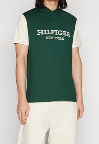 Mann in dunkelgrünem Poloshirt mit weißen Ärmeln und dem Text "Hilfiger New York", kombiniert mit hellbeigen Hosen.