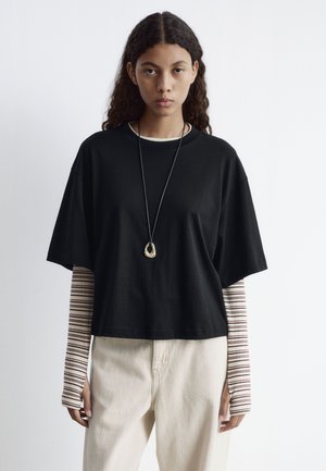 Schwarzes, kurz geschnittenes T-Shirt aus Baumwolle mit rundem Ausschnitt, getragen über einem gestreiften Langarmshirt; kombiniert mit hellbeigen Hosen.