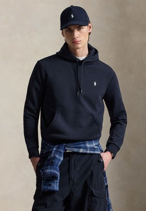 Giovane uomo che indossa una felpa con cappuccio blu navy e un cappellino con logo bianco, mani nelle tasche, camicia a quadri legata in vita, in piedi davanti a uno sfondo semplice.