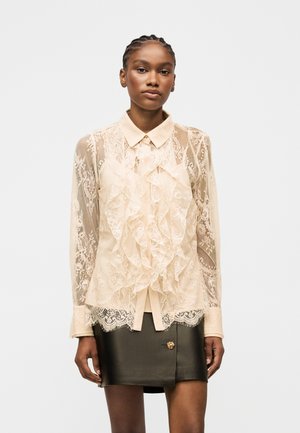 Kvinde med flettet hår iført en beige blonderusket bluse og en sort lædermininederdel med en guldknap, stående mod en hvid baggrund.